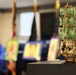 A detailed wooden model sailing ship with green-painted sails is displayed in sharp focus on a dark platform in the foreground. The ship appears to be a laser-cut wooden puzzle model, possibly resembling a fantasy or pirate-style vessel. In the blurred background, colorful artwork pieces are displayed on chairs or easels in what appears to be an indoor exhibition or display event.