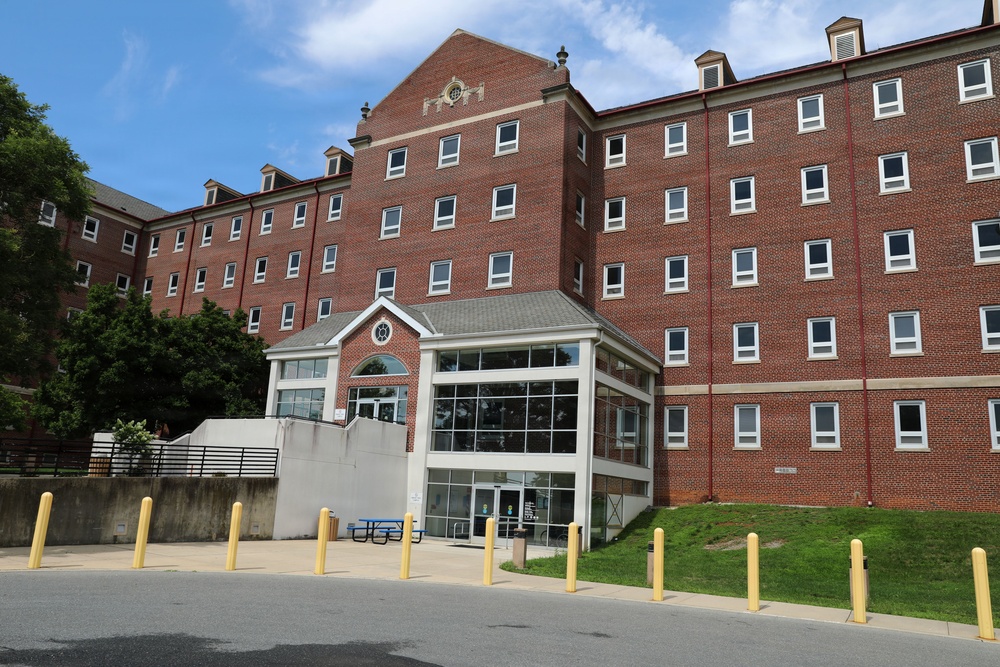 The image shows a large multi-story red brick institutional building, likely a university dormitory or academic hall, featuring a modern glass-enclosed entrance addition at ground level. The building appears to be 5-6 stories tall with a symmetrical facade, decorative gabled roofline, and uniform window placement. Yellow safety bollards line the paved area in the foreground, and well-maintained green lawns are visible.