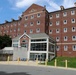 The image shows a large multi-story red brick institutional building, likely a university dormitory or academic hall, featuring a modern glass-enclosed entrance addition at ground level. The building appears to be 5-6 stories tall with a symmetrical facade, decorative gabled roofline, and uniform window placement. Yellow safety bollards line the paved area in the foreground, and well-maintained green lawns are visible.