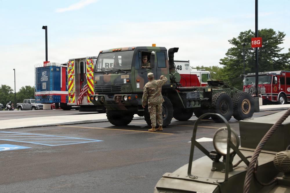 A military vehicle staging area in a parking lot, where a soldier in camouflage uniform is directing a large military tactical truck (appears to be an LMTV or FMTV). Several emergency and support vehicles are visible in the background, including fire trucks numbered 48 and an ambulance-style 'Vet Center' vehicle decorated with an American flag. The scene appears to be a veterans event or military display, indicated by the 'Vet Center' signage and organized vehicle arrangement.