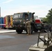 A military vehicle staging area in a parking lot, where a soldier in camouflage uniform is directing a large military tactical truck (appears to be an LMTV or FMTV). Several emergency and support vehicles are visible in the background, including fire trucks numbered 48 and an ambulance-style 'Vet Center' vehicle decorated with an American flag. The scene appears to be a veterans event or military display, indicated by the 'Vet Center' signage and organized vehicle arrangement.