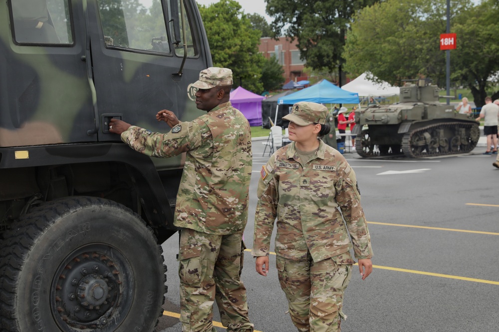 Two U.S. Army soldiers in OCP (Operational Camouflage Pattern) uniforms are inspecting or interacting with a military tactical truck at what appears to be a public military display or open house event. A vintage tank is visible in the background alongside civilian tents and bystanders. The female soldier's name tape reads 'GERNAJTAITIS' and both wear U.S. Army insignia.