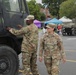Two U.S. Army soldiers in OCP (Operational Camouflage Pattern) uniforms are inspecting or interacting with a military tactical truck at what appears to be a public military display or open house event. A vintage tank is visible in the background alongside civilian tents and bystanders. The female soldier's name tape reads 'GERNAJTAITIS' and both wear U.S. Army insignia.