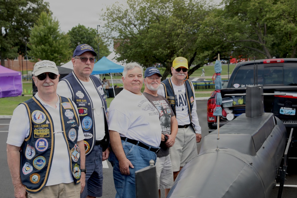 Five older men, several wearing navy blue vests adorned with military patches and pins, pose together at what appears to be an outdoor veterans event or community gathering. A gray scale model of a submarine is prominently displayed in the foreground, suggesting these men are submarine veterans. Colorful event tents and a pickup truck are visible in the background.