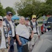 Five older men, several wearing navy blue vests adorned with military patches and pins, pose together at what appears to be an outdoor veterans event or community gathering. A gray scale model of a submarine is prominently displayed in the foreground, suggesting these men are submarine veterans. Colorful event tents and a pickup truck are visible in the background.