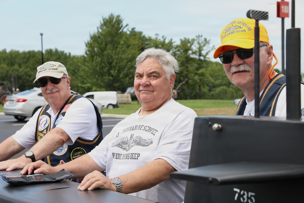 Three older men, appearing to be U.S. Navy submarine veterans, are seated together at what looks like an outdoor event or display. The men are wearing submarine service-related apparel including vests with patches, caps labeled 'Submarine Veteran,' and a t-shirt reading 'U.S. Submarine Service – Pride Runs Deep.' A model or replica submarine with the number 735 is visible in the foreground.