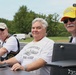 Three older men, appearing to be U.S. Navy submarine veterans, are seated together at what looks like an outdoor event or display. The men are wearing submarine service-related apparel including vests with patches, caps labeled 'Submarine Veteran,' and a t-shirt reading 'U.S. Submarine Service – Pride Runs Deep.' A model or replica submarine with the number 735 is visible in the foreground.