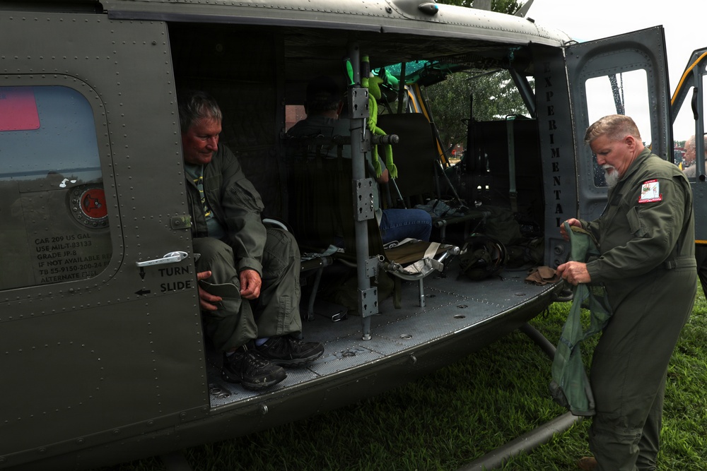 Two men wearing olive green flight suits are working around a military helicopter parked on grass, likely at an airshow or public display event. One man is seated inside the open cargo bay of the helicopter, while the other stands outside handling equipment. The helicopter appears to be a vintage military utility helicopter, possibly a UH-1 'Huey', marked with 'EXPERIMENTA' text on the fuselage.