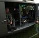 Two men wearing olive green flight suits are working around a military helicopter parked on grass, likely at an airshow or public display event. One man is seated inside the open cargo bay of the helicopter, while the other stands outside handling equipment. The helicopter appears to be a vintage military utility helicopter, possibly a UH-1 'Huey', marked with 'EXPERIMENTA' text on the fuselage.