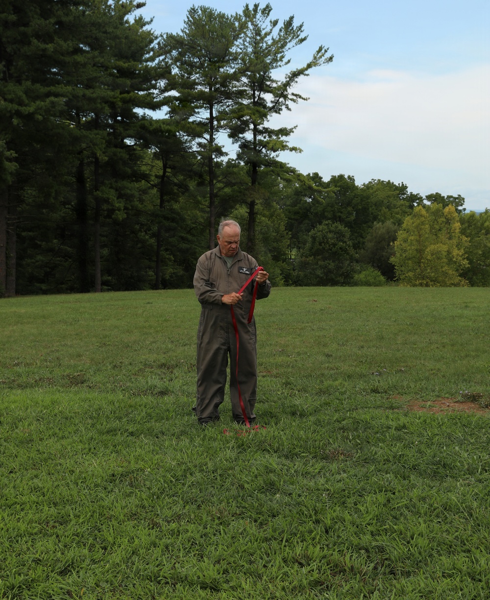An older man wearing olive/khaki coveralls stands alone in an open grassy field, handling a red strap or cord. He appears focused on the object in his hands, looking downward. The background features a treeline of tall pine and deciduous trees under a partly cloudy sky.