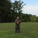 An older man wearing olive/khaki coveralls stands alone in an open grassy field, handling a red strap or cord. He appears focused on the object in his hands, looking downward. The background features a treeline of tall pine and deciduous trees under a partly cloudy sky.