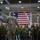 Leapfest 2026 (Pre Jump Training) A senior military officer in US Army MultiCam/OCP camouflage uniform addresses a multinational group of military personnel inside a large hangar or indoor facility. A large American flag is prominently displayed on the wall behind the assembled troops. The gathering appears to be a formal briefing or address involving personnel from multiple nations, identifiable by their varied uniforms.