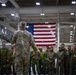 A senior military officer in US Army MultiCam/OCP camouflage uniform addresses a multinational group of military personnel inside a large hangar or indoor facility. A large American flag is prominently displayed on the wall behind the assembled troops. The gathering appears to be a formal briefing or address involving personnel from multiple nations, identifiable by their varied uniforms.