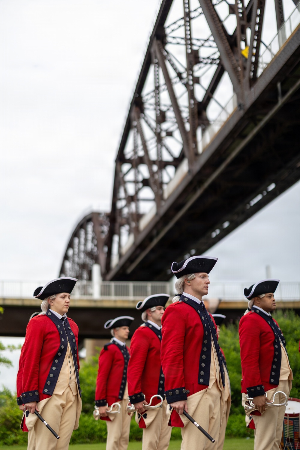 A group of performers dressed in 18th-century colonial-era military uniforms — red coats with navy trim, cream breeches, white wigs, and tricorn hats — stand in formation outdoors. They appear to be members of a ceremonial fife and drum corps, holding instruments and batons. A large steel truss bridge is visible in the background, suggesting a riverside location.