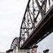 A group of performers dressed in 18th-century colonial-era military uniforms — red coats with navy trim, cream breeches, white wigs, and tricorn hats — stand in formation outdoors. They appear to be members of a ceremonial fife and drum corps, holding instruments and batons. A large steel truss bridge is visible in the background, suggesting a riverside location.