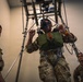 Military personnel are conducting a training exercise using a suspended harness rig inside an indoor facility. The central figure is suspended in a harness system attached to an overhead metal frame structure, wearing a head-mounted device (likely night vision goggles or VR headset) and a green chest vest. Two other soldiers in camouflage uniforms observe and assist with the training exercise.