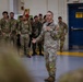 A U.S. Army officer is addressing a group of multinational military personnel in what appears to be a hangar or large indoor military facility. The speaker is holding a microphone and gesturing while speaking to a crowd of soldiers wearing various camouflage uniforms. The setting suggests a briefing, ceremony, or multinational training event.