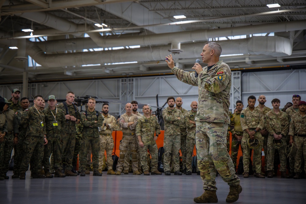 A senior military officer in US Army camouflage uniform addresses a large group of multinational military personnel inside what appears to be a military hangar or large indoor facility. The speaker is holding a microphone and gesturing with both hands, suggesting a formal briefing or address. The assembled personnel are wearing various camouflage patterns, indicating a multinational or joint military exercise or event.