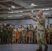 A senior military officer in US Army camouflage uniform addresses a large group of multinational military personnel inside what appears to be a military hangar or large indoor facility. The speaker is holding a microphone and gesturing with both hands, suggesting a formal briefing or address. The assembled personnel are wearing various camouflage patterns, indicating a multinational or joint military exercise or event.