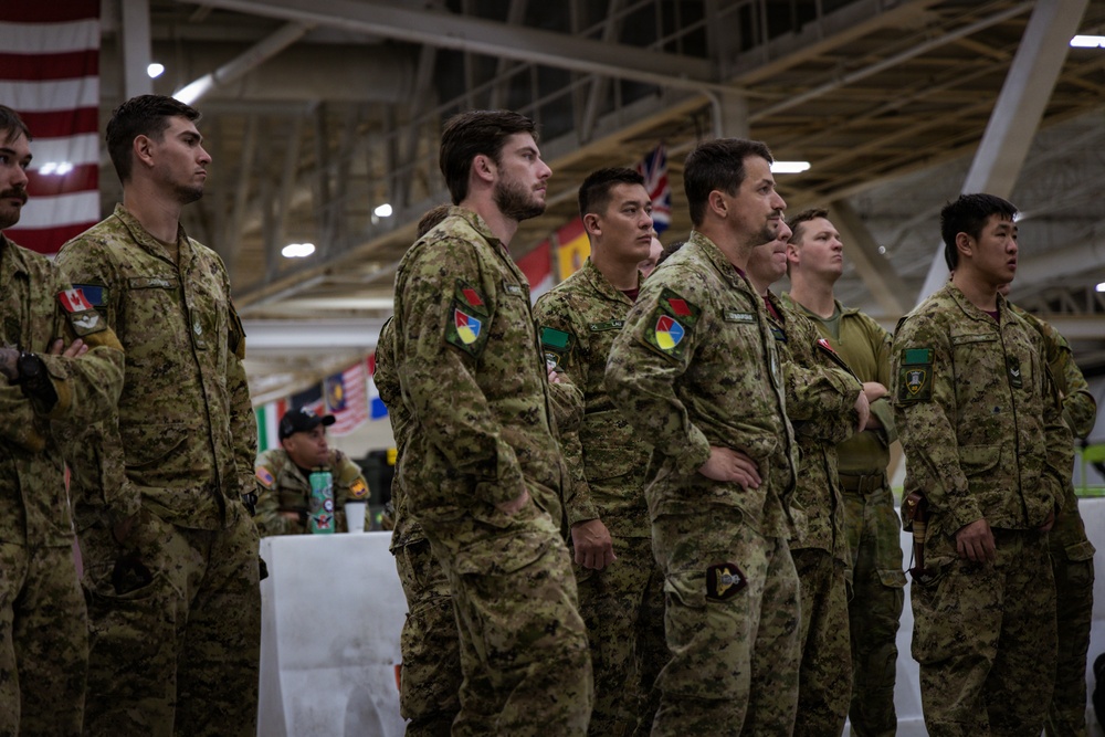 A group of military personnel in camouflage uniforms are standing at attention or in a relaxed formation inside what appears to be a large hangar or military facility. Multiple national flags are visible in the background, suggesting a multinational military event or ceremony. The soldiers display various unit patches and national flag patches on their uniforms, indicating a coalition or joint military gathering.