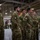 Leapfest 2026 (Pre Jump Training) A group of military personnel in camouflage uniforms are standing at attention or in a relaxed formation inside what appears to be a large hangar or military facility. Multiple national flags are visible in the background, suggesting a multinational military event or ceremony. The soldiers display various unit patches and national flag patches on their uniforms, indicating a coalition or joint military gathering.