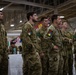 A group of military personnel in camouflage uniforms are standing at attention or in a relaxed formation inside what appears to be a large hangar or military facility. Multiple national flags are visible in the background, suggesting a multinational military event or ceremony. The soldiers display various unit patches and national flag patches on their uniforms, indicating a coalition or joint military gathering.