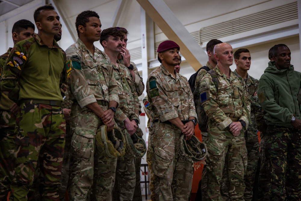 A group of military personnel from multiple nations are standing together in an indoor facility, appearing to attend a briefing or formation. The soldiers are wearing various camouflage uniforms and insignia suggesting a multinational military exercise or joint operation. One soldier is notably wearing a maroon/red beret, indicative of airborne or paratrooper units.