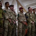 A group of military personnel from multiple nations are standing together in an indoor facility, appearing to attend a briefing or formation. The soldiers are wearing various camouflage uniforms and insignia suggesting a multinational military exercise or joint operation. One soldier is notably wearing a maroon/red beret, indicative of airborne or paratrooper units.