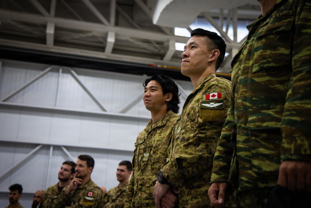 A group of military personnel standing in formation inside what appears to be a hangar or large indoor facility. The soldiers in the foreground are wearing Canadian Armed Forces CADPAT (Canadian Disruptive Pattern) uniforms, with a Canadian flag patch clearly visible on one soldier's shoulder. A soldier on the far right is wearing a different camouflage pattern, suggesting a multinational or joint military event.