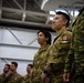 A group of military personnel standing in formation inside what appears to be a hangar or large indoor facility. The soldiers in the foreground are wearing Canadian Armed Forces CADPAT (Canadian Disruptive Pattern) uniforms, with a Canadian flag patch clearly visible on one soldier's shoulder. A soldier on the far right is wearing a different camouflage pattern, suggesting a multinational or joint military event.