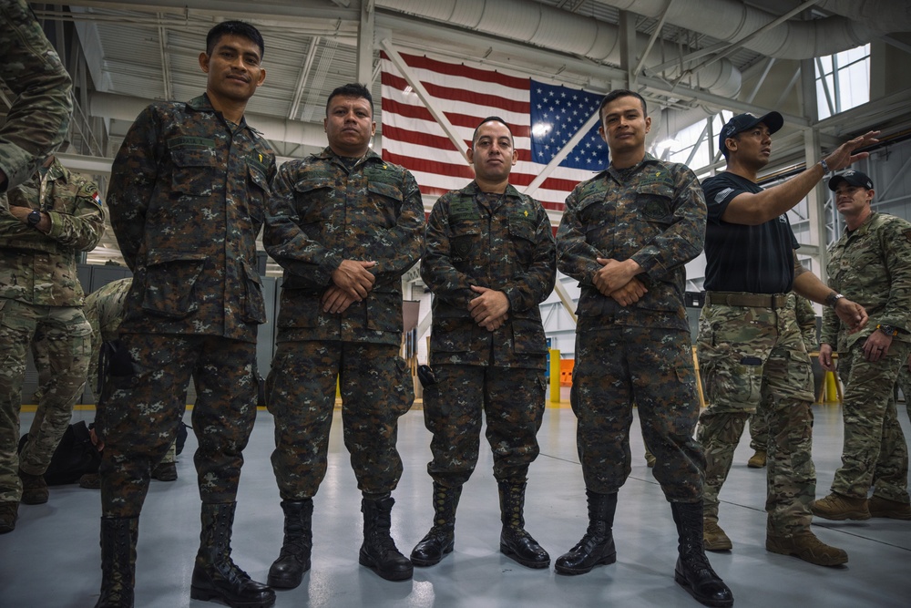 A group of military personnel from at least two different nations pose together inside what appears to be a military hangar or facility. Three soldiers in the foreground wear a distinctive woodland camouflage pattern different from the MultiCam/OCP uniforms worn by personnel on the right side. A large American flag is prominently displayed in the background, suggesting a joint military exercise or exchange program.