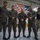 Leapfest 2026 (Pre Jump Training) A group of military personnel from at least two different nations pose together inside what appears to be a military hangar or facility. Three soldiers in the foreground wear a distinctive woodland camouflage pattern different from the MultiCam/OCP uniforms worn by personnel on the right side. A large American flag is prominently displayed in the background, suggesting a joint military exercise or exchange program.