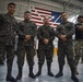 A group of military personnel from at least two different nations pose together inside what appears to be a military hangar or facility. Three soldiers in the foreground wear a distinctive woodland camouflage pattern different from the MultiCam/OCP uniforms worn by personnel on the right side. A large American flag is prominently displayed in the background, suggesting a joint military exercise or exchange program.
