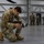 Leapfest 2026 (Pre Jump Training) A U.S. Army soldier in MultiCam/OCP camouflage uniform is kneeling on one knee on a hangar floor, adjusting or inspecting a camera equipped with a white telephoto lens and a RĂ˜DE microphone mounted on top. The soldier's name tape reads 'TOLEDO' and displays a U.S. flag patch on the right shoulder. In the background, several other soldiers in similar uniforms are gathered in small groups inside what appears to be a military aircraft hangar.
