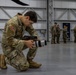 A U.S. Army soldier in MultiCam/OCP camouflage uniform is kneeling on one knee on a hangar floor, adjusting or inspecting a camera equipped with a white telephoto lens and a RØDE microphone mounted on top. The soldier's name tape reads 'TOLEDO' and displays a U.S. flag patch on the right shoulder. In the background, several other soldiers in similar uniforms are gathered in small groups inside what appears to be a military aircraft hangar.