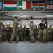 Military personnel from multiple nations are gathered inside a large hangar facility, likely during a multinational training exercise or briefing. Several soldiers in the foreground are holding yellow static lines or ropes, suggesting a parachute or airborne operations training context. The presence of international flags and diverse camouflage patterns indicates a coalition or joint military event.