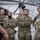 Leapfest 2026 (Pre Jump Training) A group of Canadian Armed Forces personnel are standing in a military hangar, attentively listening to a briefing or presentation. Several individuals are wearing Canadian Digital Pattern (CADPAT) camouflage uniforms with Canadian flag patches and military insignia visible. A helicopter is partially visible in the background, indicating this is likely an aviation facility.