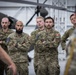 A group of Canadian Armed Forces personnel are standing in a military hangar, attentively listening to a briefing or presentation. Several individuals are wearing Canadian Digital Pattern (CADPAT) camouflage uniforms with Canadian flag patches and military insignia visible. A helicopter is partially visible in the background, indicating this is likely an aviation facility.