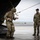Leapfest 2026 (Pre Jump Training) Two military personnel in MultiCam/OCP camouflage uniforms are positioned at the rear ramp of a military aircraft on a wet tarmac. One soldier wearing a helmet with netting is stepping off or demonstrating movement at the aircraft ramp, while a second soldier in a black cap observes. The setting appears to be a training or operational exercise at a military airfield.