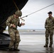 Two military personnel in MultiCam/OCP camouflage uniforms are positioned at the rear ramp of a military aircraft on a wet tarmac. One soldier wearing a helmet with netting is stepping off or demonstrating movement at the aircraft ramp, while a second soldier in a black cap observes. The setting appears to be a training or operational exercise at a military airfield.