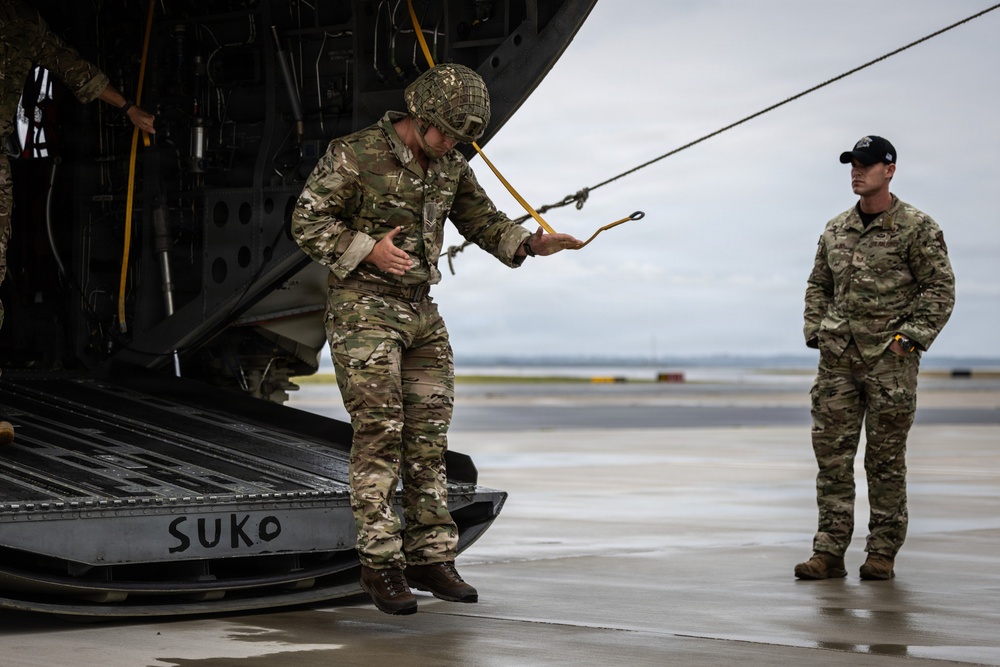 Two military personnel in MultiCam/OCP camouflage uniforms are on a wet tarmac near the open rear ramp of a large military aircraft. The central figure, wearing a helmet with netting, is handling yellow static line cables attached to the aircraft ramp. A second soldier in a black cap stands observing nearby.