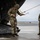 Leapfest 2026 (Pre Jump Training) Two military personnel in MultiCam/OCP camouflage uniforms are on a wet tarmac near the open rear ramp of a large military aircraft. The central figure, wearing a helmet with netting, is handling yellow static line cables attached to the aircraft ramp. A second soldier in a black cap stands observing nearby.