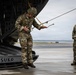 Two military personnel in MultiCam/OCP camouflage uniforms are on a wet tarmac near the open rear ramp of a large military aircraft. The central figure, wearing a helmet with netting, is handling yellow static line cables attached to the aircraft ramp. A second soldier in a black cap stands observing nearby.