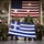 Leapfest 2026 (Pre Jump Training) Four military personnel in camouflage uniforms pose together holding a Greek national flag in what appears to be a military hangar or warehouse facility. A large United States flag is prominently displayed on the wall behind them, suggesting a joint military exercise or bilateral cooperation event between Greece and the United States. Two personnel are kneeling in front while two stand behind, holding the Greek flag horizontally for display.
