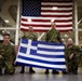 Four military personnel in camouflage uniforms pose together holding a Greek national flag in what appears to be a military hangar or warehouse facility. A large United States flag is prominently displayed on the wall behind them, suggesting a joint military exercise or bilateral cooperation event between Greece and the United States. Two personnel are kneeling in front while two stand behind, holding the Greek flag horizontally for display.