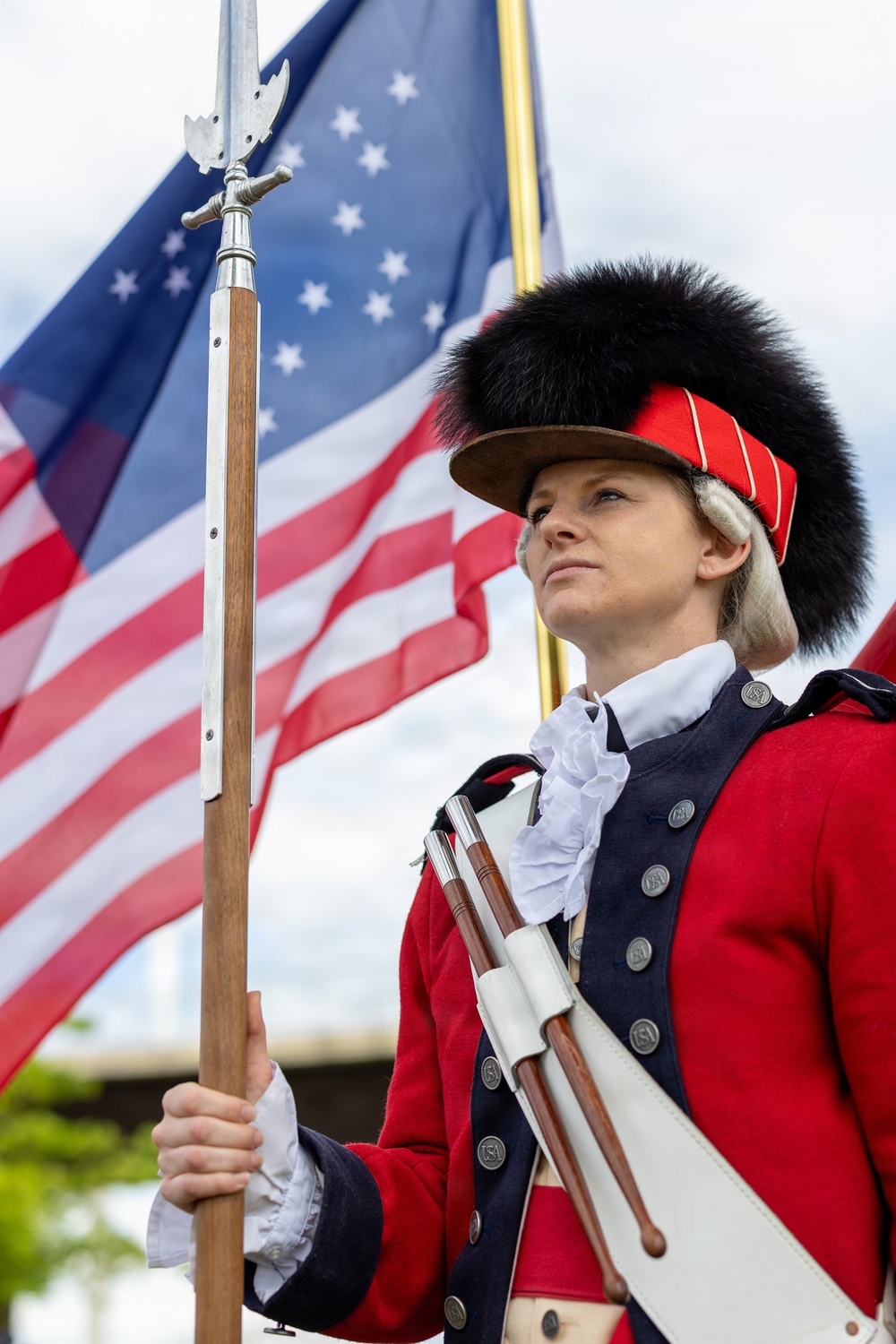 A woman dressed in an 18th-century American Revolutionary War-era military uniform stands at attention holding a flag staff with a spear-tip finial. She wears a distinctive bearskin cap with a red band, a red coat with navy blue facings, white ruffled cravat, and white cross-belt. An American flag with stars and stripes waves prominently in the background.