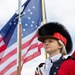 A woman dressed in an 18th-century American Revolutionary War-era military uniform stands at attention holding a flag staff with a spear-tip finial. She wears a distinctive bearskin cap with a red band, a red coat with navy blue facings, white ruffled cravat, and white cross-belt. An American flag with stars and stripes waves prominently in the background.