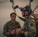 Three military personnel are engaged in what appears to be equipment fitting or training exercise. A central figure wearing British Army multicam uniform with a Union Jack patch is being fitted with a complex helmet-mounted system with wires and harnesses. A U.S. Army soldier identified by name tape as 'NORRIS' observes the procedure.
