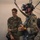 Leapfest 2026 (Pre Jump Training) Three military personnel are gathered in what appears to be an indoor training or briefing area. The central figure is wearing a British Army uniform with a Union Jack patch, equipped with night vision goggles, a parachute harness, and holding a helmet. A U.S. Army soldier with the name tape 'MORRIS' is observing on the left, while a third soldier faces away from the camera on the right.