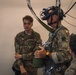 Three military personnel are gathered in what appears to be an indoor training or briefing area. The central figure is wearing a British Army uniform with a Union Jack patch, equipped with night vision goggles, a parachute harness, and holding a helmet. A U.S. Army soldier with the name tape 'MORRIS' is observing on the left, while a third soldier faces away from the camera on the right.